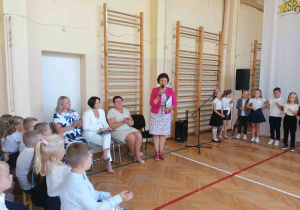 Pani mówiąca przez mikrofon, a wokół niej dzieci ubrane na galowo.