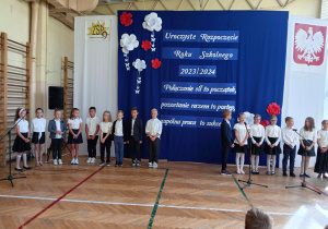 Dzieci ubrane na galowo na tle napisu: Uroczyste rozpoczęcie roku szkolnego 2023 łamane przez 2024