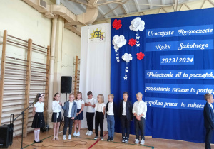 Dzieci ubrane na galowo na tle napisu: Uroczyste rozpoczęcie roku szkolnego 2023 łamane przez 2024