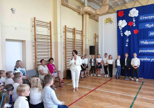 Pani mówiąca przez mikrofon, a wokół niej dzieci ubrane na galowo.