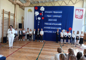 Pani mówiąca przez mikrofon, a wokół niej dzieci ubrane na galowo.