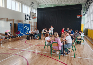 Grupy radosnych dzieci siedzą przy stolikach i piszą.