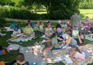 Radosne dzieci czytają książki siedząc na kocach na trawie.