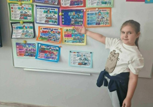 Uśmiechnięta dziewczynka wskazuje dłonią na obrazki z napisem "Tydzień bezpieczeństwa"