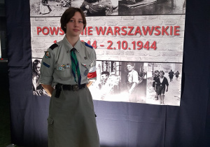 Dziewczyna w stroju harcerskim z opaską biało-czerwoną na ręce na tle plakatu z powstania warszawskiego