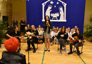 Dziewczynki z gitarami siedzące na krzesłach i dziewczyna z mikrofonem