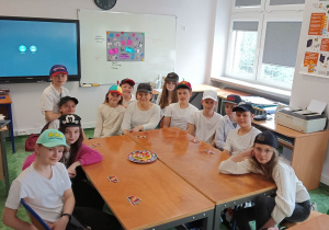 Uczniowie z panią siedzą przy stoliku ubrani na biało z czapkami na głowie