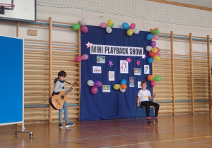 2 chłopcy, jeden z gitarą występują na środku sali
