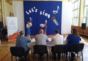 Dziewczyna i kobieta z mikrofonem na tle napisu Let's Sing oraz ludzie siedzący przy stoliku