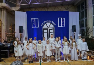 Dzieci ubrane na biało stoją w dwóch rzędach i śpiewają