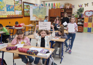 Dzieci w ławkach i dziewczynka z napisem "dzień języka ojczystego"
