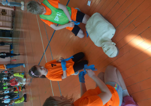 Dzieci w pomarańczowych koszulkach klęczą przy manekinie i zakładają rękawiczki jednorazowe.