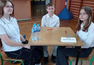 2 dziewczynki i chłopiec siedzą przy stoliku z napisem Szkoła Podstawowa nr9