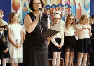Pani mówi przez mikrofon na uroczystości, za nią uśmiechnięte dzieci