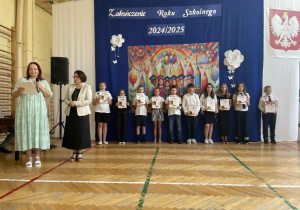 Pani mówi przez mikrofon, uśmiechnięte dzieci z nagrodami, stoją w rzędzie
