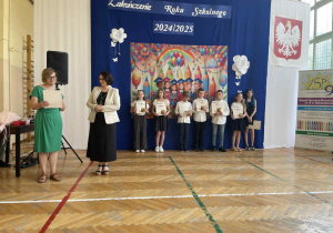 Pani mówi przez mikrofon, uśmiechnięte dzieci z nagrodami, stoją w rzędzie