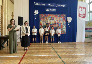 Pani mówi przez mikrofon, uśmiechnięte dzieci z nagrodami, stoją w rzędzie