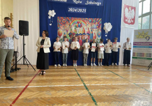 Pan mówi przez mikrofon, uśmiechnięte dzieci z nagrodami, stoją w rzędzie