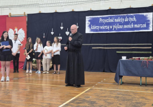 Mężczyzna w sutannie i uczniowie na tle granatowej dekoracji