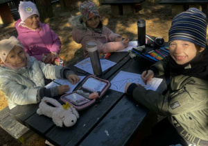 Uśmiechnięte dzieci siedzą przy stoliku w lesie i kolorują obrazek