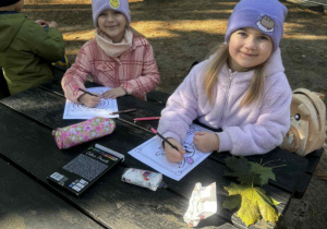Uśmiechnięte dziewczynki siedzą przy stoliku w lesie i kolorują obrazek