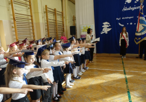 Radosne dzieci ubrane odświętnie stoją w rzędzie z wyciągniętymi prawymi dłońmi