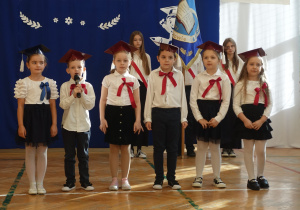 Dzieci ubrane na galowo z biretami na głowach stoją w rzędzie