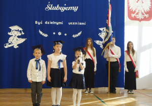 Dzieci ubrane na galowo z biretami na głowach stoją w rzędzie