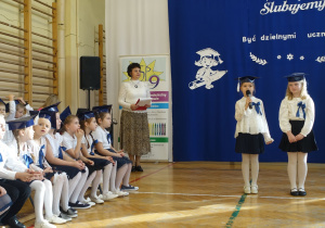 Dzieci ubrane na galowo z biretami na głowach stoją w rzędzie