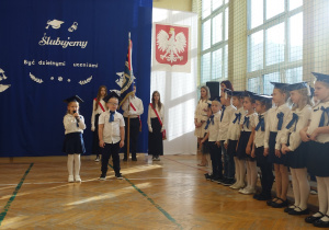 Dzieci ubrane na galowo z biretami na głowach stoją w rzędzie