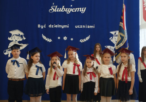 Dzieci ubrane na galowo z biretami na głowach stoją w rzędzie