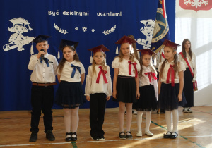 Dzieci ubrane na galowo z biretami na głowach stoją w rzędzie