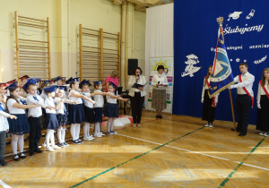 Radosne dzieci ubrane odświętnie stoją w rzędzie z wyciągniętymi prawymi dłońmi