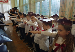 Radosne dzieci ubrane odświętnie stoją w rzędzie z wyciągniętymi prawymi dłońmi