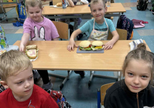 Uśmiechnięte dzieci siedzą przy stoliku a przed nimi kolorowe kanapki