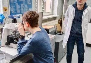 Mężczyzna w białym fartuchu, obok chłopak ogląda coś przeze mikroskop