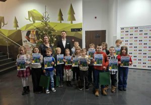 Grupa uśmiechniętych dzieci z dorosłymi pozują do zdjęcia z torbą i dyplomem w dłoniach