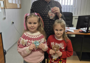 Kobieta trzyma piernik, przed nią stoją dziewczynki, które trzymają pierniczki