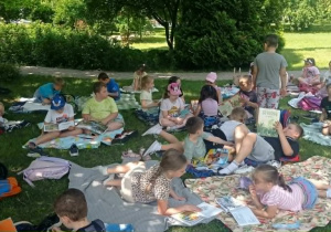 Radosne dzieci czytają książki siedząc na kocach na trawie.