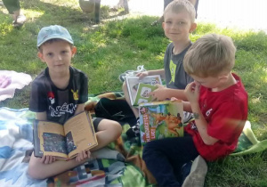Radośni chłopcy czytają książki siedząc na kocach na trawie.