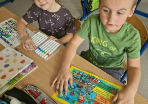 Chłopiec z dziewczynką siedzą w ławce a przed nimi obrazki z napisem "Tydzień bezpieczeństwa"