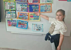Uśmiechnięta dziewczynka wskazuje dłonią na obrazki z napisem "Tydzień bezpieczeństwa"