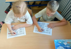 Chłopiec z dziewczynką siedzą w ławce a przed nimi obrazki z napisem "Tydzień bezpieczeństwa"