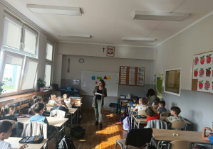 Kobieta na środku sali mówi do dzieci siedzących w ławkach