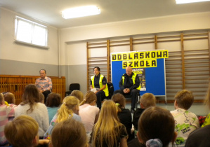 Mężczyzna z policjantami w mundurach, ubrani w kamizelki odblaskowe siedzą przed grupą uczniów