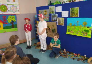 Dzieci na spotkaniu z panią leśnik mówią wiersz w grzybowych czapkach na głowie
